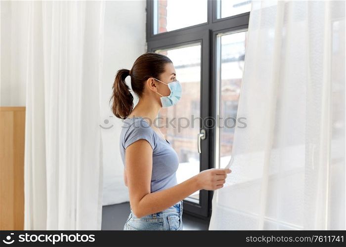 health, safety and pandemic concept - sick young woman wearing protective medical mask looking through window at home. sick young woman wearing protective medical mask