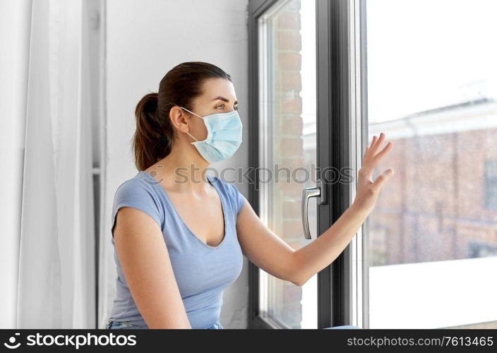 health, safety and pandemic concept - sick young woman wearing protective medical mask looking through window at home. sick young woman wearing protective medical mask