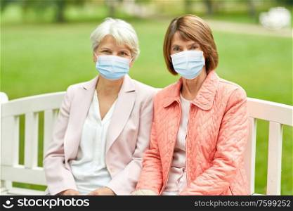 health, safety and pandemic concept - two senior women or friends in medical masks for protection from virus sitting on bench at summer park. senior women in medical masks at park