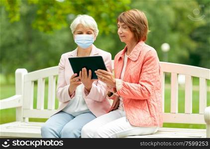 health, safety and pandemic concept - two senior women or friends in medical mask for protection from virus tablet pc computer sitting on bench at summer park. old women in medical mask with tablet pc at park