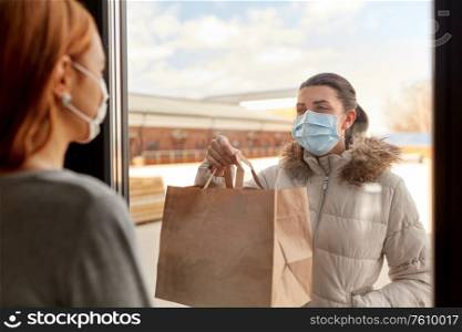 health, safety and pandemic concept - young woman wearing protective medical mask with food in paper bag looking through window at her friend staying at home. woman in medical mask delivers food for her friend