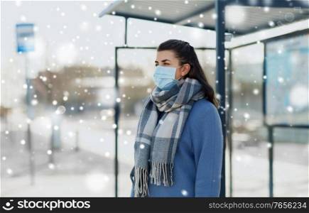 health, safety and pandemic concept - young woman wearing protective medical mask at bus stop in city over snow. young woman wearing medical mask at bus stop