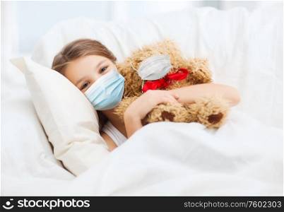 health safety, quarantine and pandemic concept - girl in protective medical mask with teddy bear in bed. girl wearing medical mask with teddy bear in bed