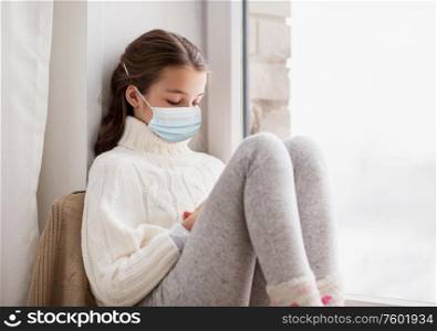 health safety, quarantine and pandemic concept - sad beautiful girl in protective medical mask for protection from virus disease sitting on sill at home window in winter. sad girl in medical mask sitting on sill at home