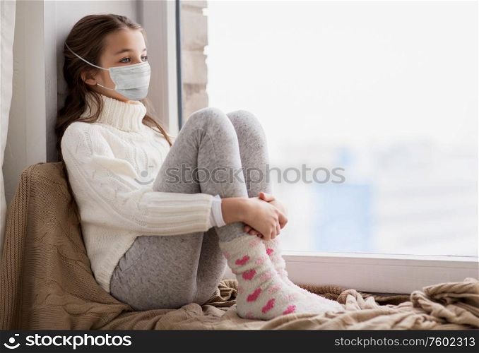 health safety, quarantine and pandemic concept - sad beautiful girl in protective medical mask for protection from virus disease sitting on sill at home window in winter. sad girl in medical mask sitting on sill at home