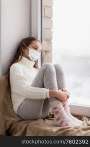 health safety, quarantine and pandemic concept - sad beautiful girl in protective medical mask for protection from virus disease sitting on sill at home window in winter. sad girl in medical mask sitting on sill at home