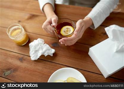 health, traditional medicine and ethnoscience concept - close up of ill woman drinking tea with lemon, honey, ginger and used paper tissue on wooden table