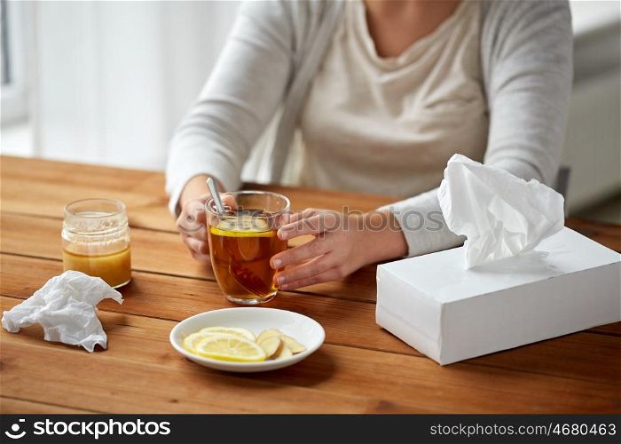 health, traditional medicine and ethnoscience concept - close up of ill woman drinking tea with lemon, honey and ginger at wooden table