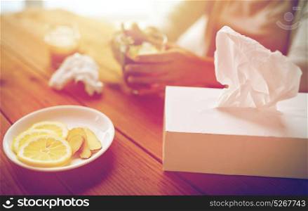 health, traditional medicine and ethnoscience concept - ill woman drinking tea with lemon, honey and ginger and paper wipes box on wooden table. ill woman drinking tea with lemon and ginger