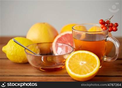 health, traditional medicine, folk remedy and ethnoscience concept - cup of tea with honey, lemon and rowanberry on wooden background