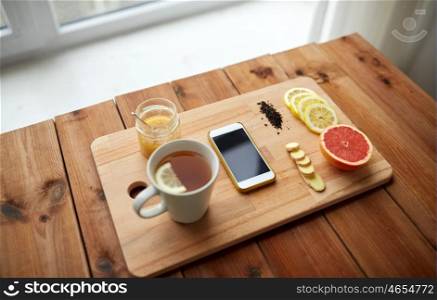 health, traditional medicine, folk remedy and ethnoscience concept - smartphone with cup of ginger tea, honey and citrus on wooden board