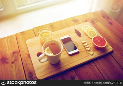 health, traditional medicine, folk remedy and ethnoscience concept - smartphone with cup of ginger tea, honey and citrus on wooden board. smartphone with cup of lemon tea, honey and ginger