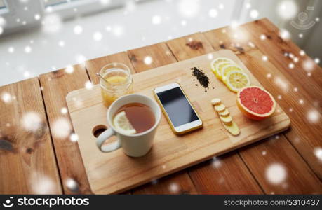 health, traditional medicine, folk remedy and ethnoscience concept - smartphone with cup of ginger tea, honey and citrus on wooden board over snow. smartphone with cup of lemon tea, honey and ginger