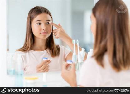 health, vision and old people concept - teenage girl applying contact lenses in front of mirror at home bathroom. teenage girl applying contact lenses at bathroom