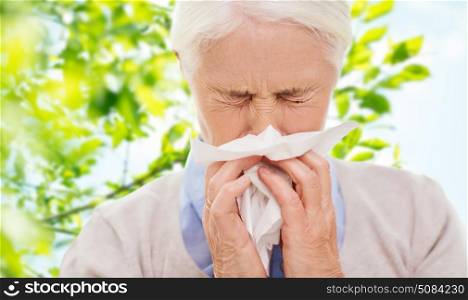 healthcare, age and people concept - sick senior woman blowing nose to paper napkin over green natural background. sick senior woman blowing nose to paper napkin. sick senior woman blowing nose to paper napkin