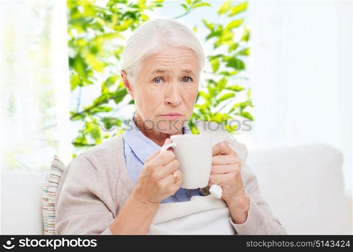 healthcare, age and people concept - sick senior woman with paper napkin drinking hot tea at home over window with green natural background. sick senior woman drinking hot tea at home