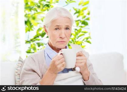 healthcare, age and people concept - sick senior woman with paper napkin drinking hot tea at home over window with green natural background. sick senior woman drinking hot tea at home