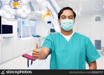healthcare and medicine concept - doctor, surgeon or male nurse in blue uniform and medical mask showing thumbs up over surgery ward at hospital on background. male doctor in mask showing thumbs up at clinic