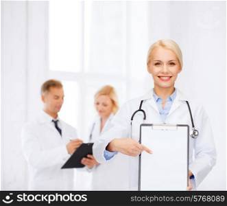healthcare and medicine concept - smiling female doctor with stethoscope pointing finger to clipboard and blank page