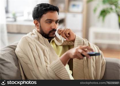 healthcare, cold, hygiene and people concept - sick indian man in blanket with paper tissue and tv remote at home. sick man with paper tissue and tv remote at home