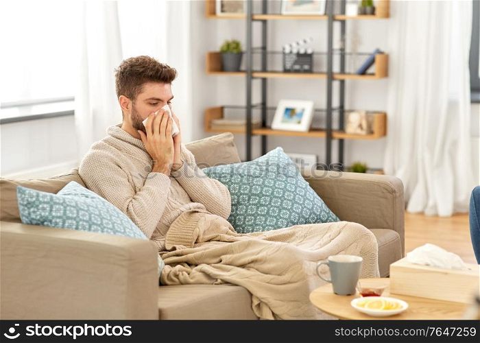 healthcare, cold, hygiene and people concept - sick man in blanket with paper tissue blowing his nose at home. sick man blowing nose in paper tissue at home