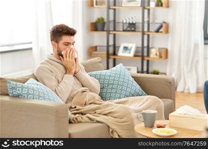 healthcare, cold, hygiene and people concept - sick man in blanket with paper tissue blowing his nose at home. sick man blowing nose in paper tissue at home
