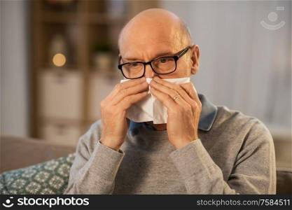 healthcare, flu and people concept - sick senior man with paper tissue blowing his nose at home in evening. sick senior man with paper tissue blowing his nose