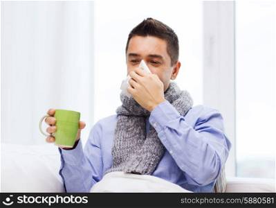 healthcare, flu, people, rhinitis and medicine concept - ill man blowing his nose with paper napkin and drinking tea at home
