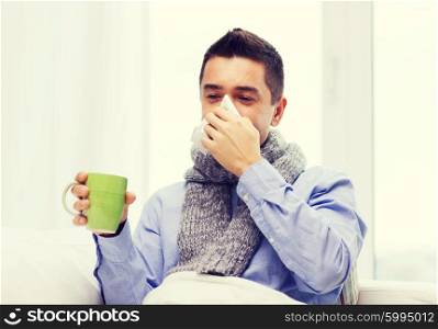 healthcare, flu, people, rhinitis and medicine concept - ill man blowing his nose with paper napkin and drinking tea at home