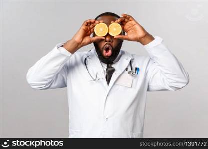 Healthcare, healthy diet and disease concept. Funny good-looking physician, african-american doctor playing with orange, look amused, tell stay safe, eat vitamins and fruits, grey background.. Healthcare, healthy diet and disease concept. Funny good-looking physician, african-american doctor playing with orange, look amused, tell stay safe, eat vitamins and fruits, grey background