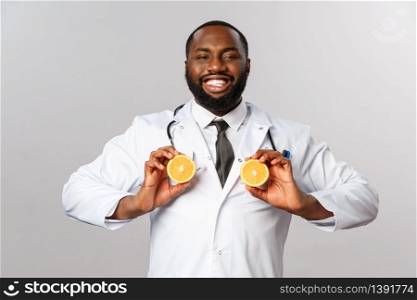 Healthcare, healthy diet and disease concept. Funny handsome african-american male doctor, physician smiling pleased, holding fruit like breast, fool around with orange, stand grey background.. Healthcare, healthy diet and disease concept. Funny handsome african-american male doctor, physician smiling pleased, holding fruit like breast, fool around with orange, stand grey background