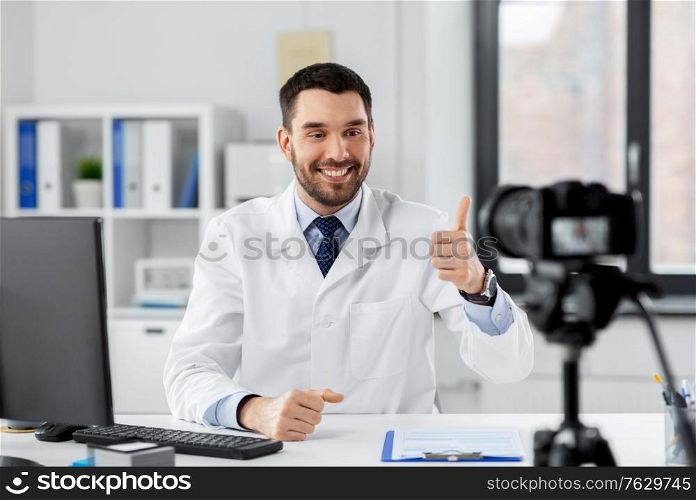 healthcare, medicine and blogging concept - happy smiling male doctor with camera recording video blog at hospital and showing thumbs up. male doctor recording video blog at hospital