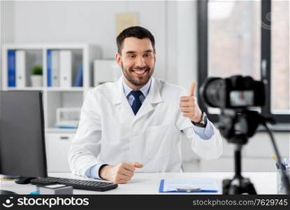 healthcare, medicine and blogging concept - happy smiling male doctor with camera recording video blog at hospital and showing thumbs up. male doctor recording video blog at hospital
