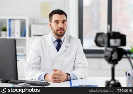 healthcare, medicine and blogging concept - male doctor with camera recording video blog at hospital. male doctor recording video blog at hospital