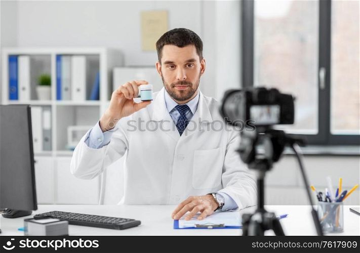 healthcare, medicine and blogging concept - male doctor with camera showing drug and recording video blog at hospital. doctor with drug recording video blog at hospital