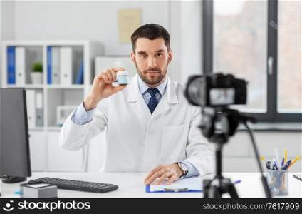 healthcare, medicine and blogging concept - male doctor with camera showing drug and recording video blog at hospital. doctor with drug recording video blog at hospital