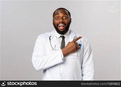 Healthcare, medicine and epidemic concept. Cheerful happy, smiling african-american doctor inviting new clients to his clinic, grinning and looking camera enthusiastic, pointing finger right corner.. Healthcare, medicine and epidemic concept. Cheerful happy, smiling african-american doctor inviting new clients to his clinic, grinning and looking camera enthusiastic, pointing finger right corner