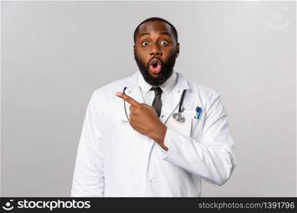 Healthcare, medicine and epidemic concept. Impressed african-american male doctor look with interest at camera as discuss new pills or medication, pointing fingers upper left corner.. Healthcare, medicine and epidemic concept. Impressed african-american male doctor look with interest at camera as discuss new pills or medication, pointing fingers upper left corner