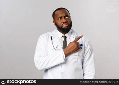 Healthcare, medicine and epidemic concept. Skeptical and displeased african-american doctor, male physician look and pointing upper right corner, grimacing at something bad, grey background.. Healthcare, medicine and epidemic concept. Skeptical and displeased african-american doctor, male physician look and pointing upper right corner, grimacing at something bad, grey background
