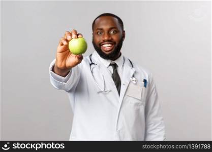 Healthcare, medicine and healthy lifestyle concept. Apple a day. Cheerful handsome african-american doctor giving patient green apple, showing fruit ask eat fruits and vitamins, grey background.. Healthcare, medicine and healthy lifestyle concept. Apple a day. Cheerful handsome african-american doctor giving patient green apple, showing fruit ask eat fruits and vitamins, grey background