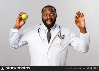Healthcare, medicine and healthy lifestyle concept. Enthusiastic african-american doctor advice stay healthy with eating more vitamins, fruits, hold apple and pills, standing grey background.. Healthcare, medicine and healthy lifestyle concept. Enthusiastic african-american doctor advice stay healthy with eating more vitamins, fruits, hold apple and pills, standing grey background