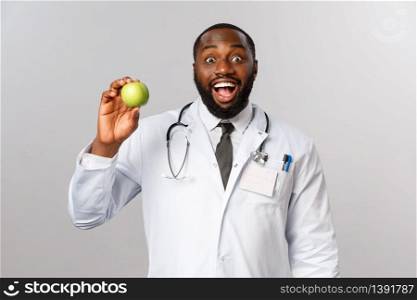 Healthcare, medicine and healthy lifestyle concept. Enthusiastic, amused happy african-american doctor advice eat fruits, showing apple and smiling, persuade kid eat vitamins, grey background.. Healthcare, medicine and healthy lifestyle concept. Enthusiastic, amused happy african-american doctor advice eat fruits, showing apple and smiling, persuade kid eat vitamins, grey background