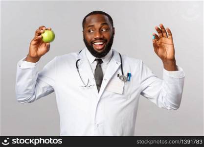Healthcare, medicine and healthy lifestyle concept. Friendly african-american doctor in white coat, holding apple and pill, suggest patients stay healthy without medication, eat more fruits.. Healthcare, medicine and healthy lifestyle concept. Friendly african-american doctor in white coat, holding apple and pill, suggest patients stay healthy without medication, eat more fruits