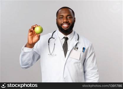 Healthcare, medicine and healthy lifestyle concept. Good-looking smiling african-american doctor advice stay fit and eat more fruits consist vitamins, showing apple to patient, grey background.. Healthcare, medicine and healthy lifestyle concept. Good-looking smiling african-american doctor advice stay fit and eat more fruits consist vitamins, showing apple to patient, grey background