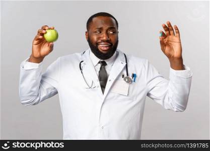 Healthcare, medicine and healthy lifestyle concept. Skeptical handsome african-american doctor look unsure and hesitant and pill as showing drugs and apple, healthy diet or medication.. Healthcare, medicine and healthy lifestyle concept. Skeptical handsome african-american doctor look unsure and hesitant and pill as showing drugs and apple, healthy diet or medication