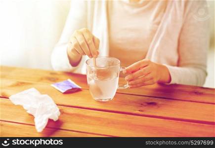 healthcare, medicine and people concept - close up of woman stirring medication in cup with spoon and paper tissue on wooden table. woman stirring medication in cup with spoon