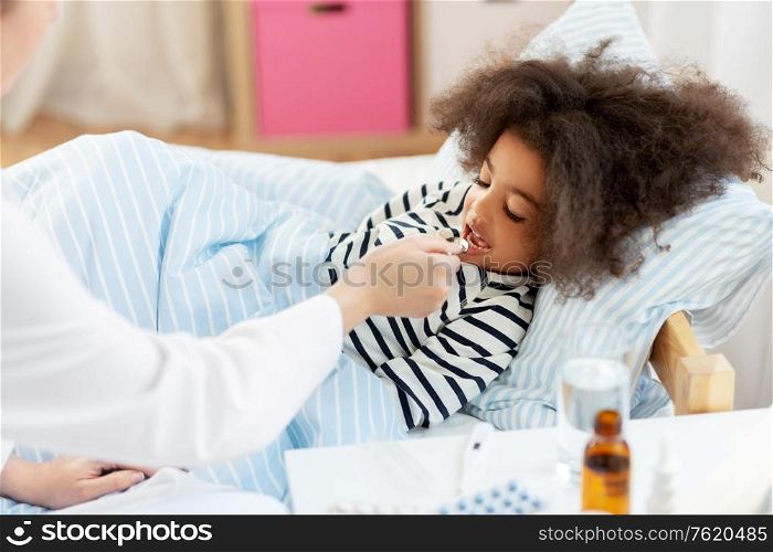 healthcare, medicine and people concept - doctor giving cough syrup to little sick african american girl in bed at home. doctor giving medicine to sick girl in bed at home