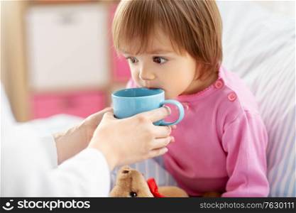 healthcare, medicine and people concept - doctor giving hot tea to little sick girl at home. doctor giving hot tea to sick little girl in bed