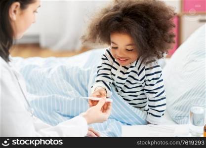 healthcare, medicine and people concept - doctor showing thermometer to happy smiling sick little african american girl in bed at home. doctor showing thermometer to smiling sick girl