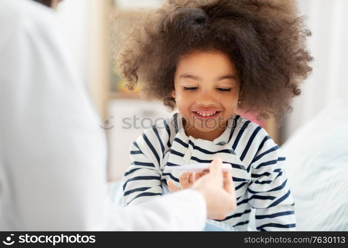 healthcare, medicine and people concept - doctor showing thermometer to happy smiling sick little african american girl in bed at home. doctor showing thermometer to smiling sick girl